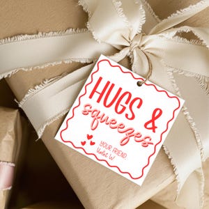 May include: A wrapped gift with a cream-colored ribbon and a gift tag that reads "Hugs & Squeezes." The tag has a red border and the words are in red and pink. The gift is wrapped in brown paper.