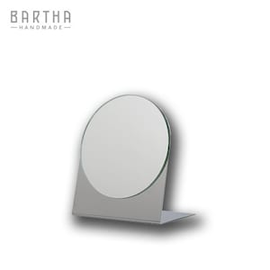 Può includere: Uno specchio da tavolo color argento con una superficie riflettente rotonda. Lo specchio è su un supporto con una base triangolare.