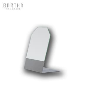 Può includere: Un moderno ed essenziale espositore in metallo con un design rettangolare e angolato. L'espositore presenta una superficie bianca e una base grigia. Il testo "BARTHA - HANDMADE-" è visibile in alto a sinistra.