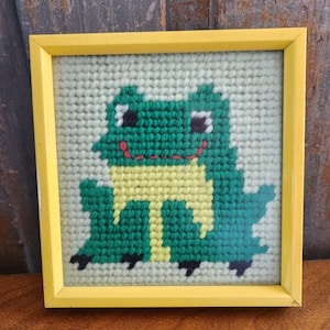 May include: A framed green and yellow frog cross-stitch with black eyes and a red mouth. The frog is sitting on a white background.