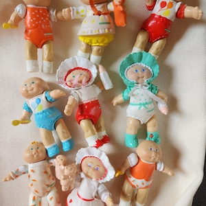 May include: A collection of vintage Cabbage Patch Kids dolls. The dolls are dressed in various outfits, including rompers, dresses, and bonnets. Some dolls are holding accessories like bottles and small dolls. The dolls have the signature Cabbage Patch Kids faces.