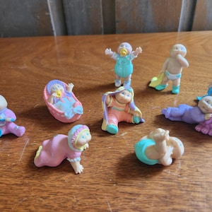 May include: A collection of small, colorful baby figurines in various poses. The figurines are in pastel colors, including pink, blue, and yellow. Some are wearing diapers or outfits, while others are in a cradle or crawling.