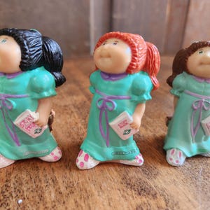 May include: Three Cabbage Patch Kids figurines. Each doll wears a teal dress with a purple bow and holds a book. The dolls have different hair colors: black, red, and brown. The dolls' feet are white with pink polka dots.