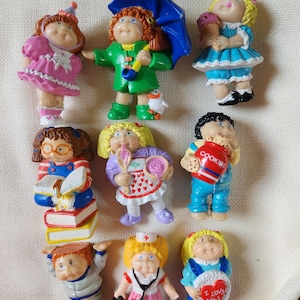 May include: A collection of vintage Cabbage Patch Kids figurines. The dolls are dressed in various outfits, including a nurse's uniform, a chef's apron, and a blue raincoat with an umbrella. Some dolls are holding accessories like books, cookies, and ice cream.