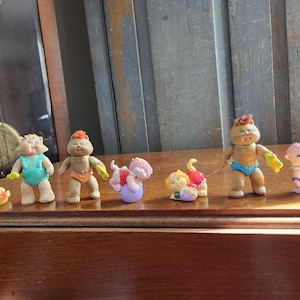 May include: A collection of vintage Cabbage Patch Kids figurines. The figures are in various poses and outfits, including swimsuits and other colorful attire. The figurines are displayed on a wooden surface.