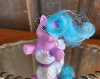 May include: A purple My Little Pony with a white and iridescent mermaid tail and blue hair. The pony is standing on a mirrored surface.