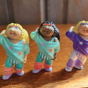 May include: Three vintage Cabbage Patch Kids figurines in athletic outfits. The figures have blonde, black, and yellow hair, and wear colorful outfits with star accents. The figurines are posed in a dynamic, athletic stance.