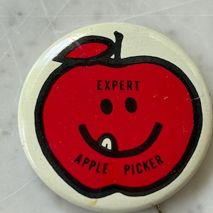 May include: A round pinback button with a cream-colored background and a red apple illustration in the center. The apple features a black smiley face with a single tooth and the text "EXPERT APPLE PICKER" printed in black capital letters.