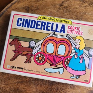 May include: A vintage box of Cinderella cookie cutters. The box features a colorful illustration of Cinderella's carriage, a horse, and Cinderella in a blue dress. The text "CINDERELLA COOKIE CUTTERS" is prominently displayed.
