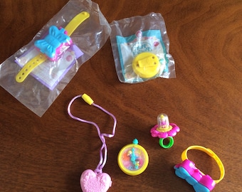 May include: A collection of five colorful plastic toys, including a butterfly-shaped bracelet, a heart-shaped locket, a ring with a flower and a small figure inside, a yellow watch with a clear face, and a yellow and blue bracelet with a flower design. The toys are all packaged in clear plastic bags.