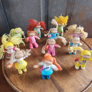 May include: A collection of small, vintage dolls with yarn hair and colorful outfits. The dolls are posed on a wooden tray, showcasing their various outfits in shades of pink, blue, yellow, and green. The dolls have painted faces and are in a playful pose.
