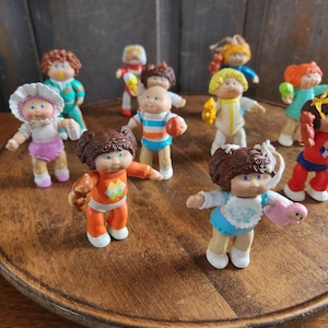 May include: A collection of vintage Cabbage Patch Kids figurines. The dolls have various hair colors, outfits, and accessories. They are displayed on a wooden serving tray. The dolls are in various poses, some holding toys.