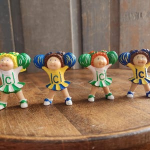 May include: Four small figurines of cheerleaders with different colored hair and outfits. Each figurine has a smiling face and is wearing a cheerleader uniform with the letter "C" on the front. The figurines are standing on a wooden surface.