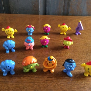 May include: A collection of 13 small plastic figurines of Mr. Men characters. The characters are all different colors and have different facial expressions. They are all standing on a wooden surface.