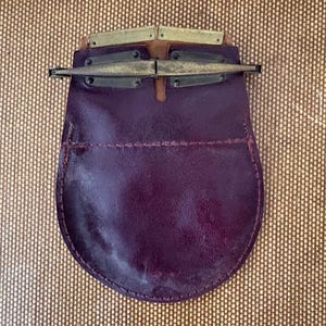 May include: A dark purple leather sporran featuring a rounded bottom shape and a horizontal stitched seam across the middle. The top closure is secured with a metallic brass-toned hardware mechanism consisting of two hinged metal plates.