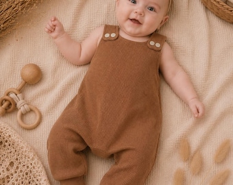 Op de afbeelding: Een lachende baby in een bruine wafel gebreide romper met knoopsluitingen op de schouders. De baby ligt op een beige deken, met een houten rammelaar en gedroogde planten in de buurt. Een beige hoofdband met strik siert het hoofd van de baby.