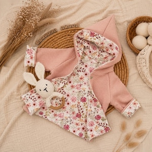 May include: A pink baby jacket with a floral print lining and hood, alongside a white crochet bunny toy with a wooden ring. The jacket is made of a textured fabric. The background includes a woven basket and dried flowers.