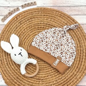 Può includere: Un sonaglio a forma di coniglio bianco all'uncinetto con un anello di legno, un cappello da neonato con stampa leopardata marrone e bianca con un bordo marrone e un'etichetta bianca che dice "Lina". Gli articoli sono su un cesto di vimini marrone.