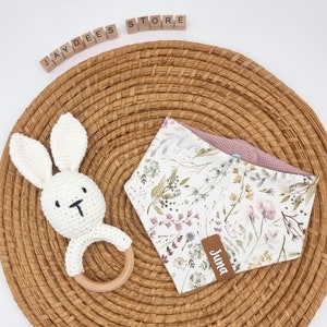 May include: A white crocheted bunny rattle with a wooden ring, a pink and white floral bandana with the name "Juna" on it, and a woven wicker basket.