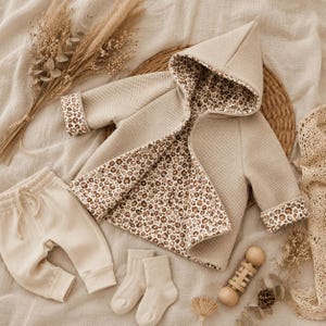 May include: A beige baby outfit set, including a hooded jacket with a leopard print lining, matching pants, and socks. A wooden rattle is also present. The set is displayed on a neutral-toned surface.