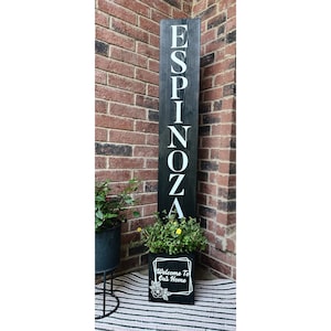 May include: Black wooden sign with the name "Espinoza" spelled vertically in white letters. The sign is in front of a brick wall and has a black planter box with a floral design and the text "Welcome to Our Home" in white letters.