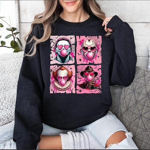 Peut inclure: Un sweat-shirt noir avec un motif graphique de quatre personnages de films d'horreur, chacun soufflant une bulle de chewing-gum rose et portant des lunettes de soleil en forme de c&oelig;ur. Le motif est sur un fond rose avec des toiles d'araign&eacute;es et des araign&eacute;es.