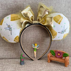 May include: A gold and black headband with large mouse ears. The ears are white with a pattern of yellow shapes and a cartoon fairy. A gold bow sits atop the headband. A small fairy figurine and miniature bench are in front.