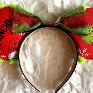 Peut inclure: Un serre-tête avec deux oreilles circulaires en tissu rouge bordées de vert pelucheux. Les oreilles présentent des flocons blancs, un personnage vert avec un bonnet de Noël et le texte Merry GRINCHMAS. Un grand nœud rayé vert et rouge brille au centre.
