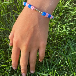 May include: A handmade bracelet featuring blue and pink disc-shaped beads, interspersed with small gold-colored beads. The bracelet is worn on a wrist, set against a backdrop of green grass.