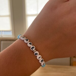May include: A beaded bracelet spelling out "CAPE GIRL" in black letters on white beads. The bracelet includes light blue accent beads and a silver-colored spacer bead. The bracelet is worn on a wrist.
