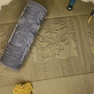 May include: A close-up of a rolling pin with a moose and pine tree design. The rolling pin is used to create an impression in clay. The clay is imprinted with the moose and pine tree design.