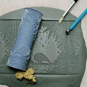 May include: A gray clay slab with a detailed kiwi bird and fern design. A cylindrical roller with a matching kiwi and fern pattern is next to the slab. A small brush and a tool with a ball tip are also present.