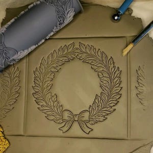 Puede incluir: Un primer plano de una baldosa de arcilla con un dise&ntilde;o en relieve de una corona con un lazo. La corona est&aacute; hecha de hojas y el lazo est&aacute; atado con una cinta. La baldosa est&aacute; rodeada por un borde de hojas.