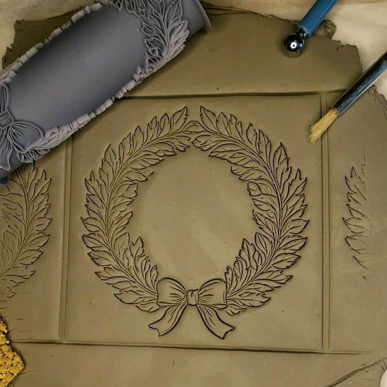 Puede incluir: Un primer plano de una baldosa de arcilla con un dise&ntilde;o en relieve de una corona con un lazo. La corona est&aacute; hecha de hojas y el lazo est&aacute; atado con una cinta. La baldosa est&aacute; rodeada por un borde de hojas.