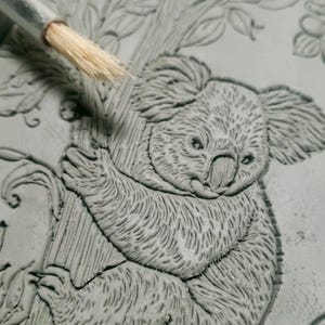 May include: Close-up of a gray koala bear sculpture clinging to a tree branch. The koala has detailed fur texture and is surrounded by carved leaves. A paintbrush is visible in the upper left corner, suggesting the sculpture is being worked on.