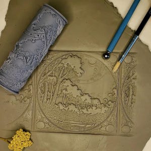 May include: A close-up of a clay tile with a detailed embossed design of a forest scene. A rolling pin with a similar design is visible in the upper left corner, along with a small tool and a paintbrush.