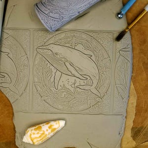 May include: A close-up of a gray clay tile with an intricate design of a whale swimming in a circle of seaweed. The tile is surrounded by other tiles with similar designs.
