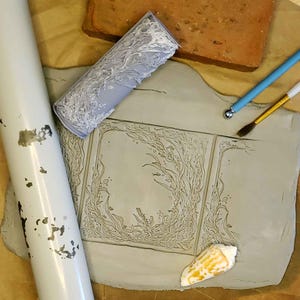 May include: A close-up of a clay tile with an intricate design of seaweed and a seashell. The tile is surrounded by tools for creating pottery, including a rolling pin, a small tool with a ball on the end, and a paintbrush.