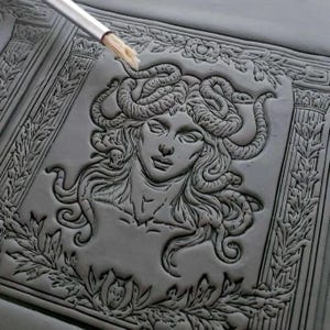 May include: A gray, textured, rectangular plaque with a detailed embossed image of Medusa, a Greek mythological figure with snakes for hair, surrounded by a decorative border of leaves and flowers.
