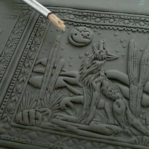 May include: A gray ceramic tile with a raised design of a coyote howling at the moon, surrounded by cacti and stars. The tile is decorated with a border of repeating geometric patterns.