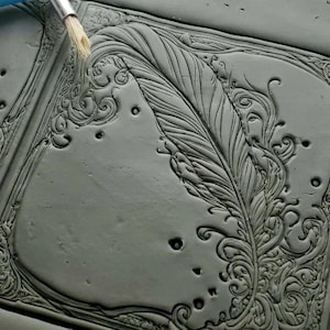 May include: A gray clay tile with a detailed, embossed design of a feather and swirling patterns. The tile is being painted with a brush.