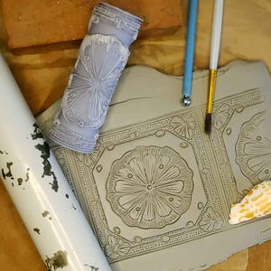 May include: A close-up of a gray clay tile with a decorative floral pattern. The tile is being used to create a decorative design. There are two tools, a blue rolling pin and a gold paintbrush, on the surface of the clay.