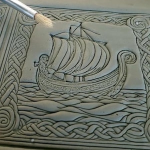 May include: A gray, rectangular ceramic tile with a detailed relief carving of a Viking ship sailing on wavy water. The tile is framed by a Celtic knot border. A paintbrush is touching the surface.