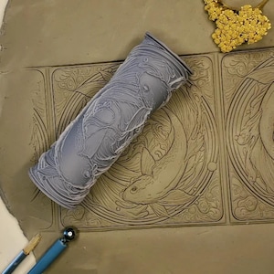 May include: A gray rolling pin with a detailed design of fish and waves. The rolling pin is on top of a clay surface with two squares that have been embossed with the same design.