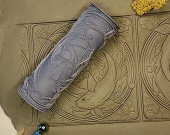 Peut inclure: Un rouleau à pâtisserie gris avec un motif détaillé de poissons et de vagues. Le rouleau à pâtisserie est posé sur une surface en argile avec deux carrés qui ont été embossés avec le même motif.