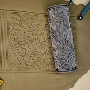 May include: A gray rolling pin with a leaf pattern design. The rolling pin is used to create an imprint of the leaf pattern on clay.