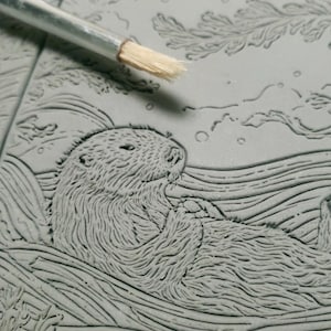 May include: A detailed carving of a sea otter resting in water, with a brush in the upper left corner. The otter is depicted with textured fur and is surrounded by carved water lines and aquatic plants. The carving is in shades of gray.