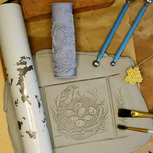 May include: A close-up of a clay tile with an embossed design of a bird in a nest with eggs. The tile is surrounded by tools for clay sculpting, including a rolling pin, a small brush, and two tools with ball-shaped ends.