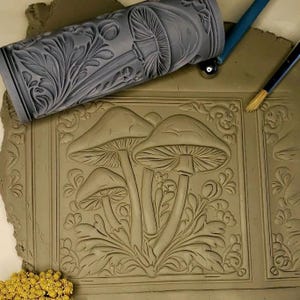 May include: A close-up of a clay tile with an embossed design of three mushrooms with stems and leaves. The tile is surrounded by a decorative border. A rolling pin with a mushroom design is visible in the background.