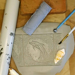 May include: A gray clay tile with an embossed design of a dolphin swimming in a circle of flowers and bubbles. The tile is surrounded by tools for creating clay art, including a rolling pin, a rubber stamp, a paintbrush, and a sculpting tool.